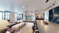 This thoughtfully designed clubroom offers a spacious, single-level layout that balances social gathering and relaxed lounging. The open floor plan is anchored by a striking floor-to-ceiling tiled fireplace, which serves as a central architectural focal point. Flanking the fireplace are two distinct seating areas: one featuring a plush olive-green sofa and a rounded armchair paired with a low coffee table, and another arranged atop a rich burgundy area rug with boucle accent chairs and a leather sofa. Light wood-look flooring extends throughout the open areas, contributing to the warm, contemporary aesthetic. A full bar zone along the back wall showcases deep teal cabinetry with arched niches, white brick backsplash, pendant lighting, and a curved island with bar seating—ideal for entertaining. Curated artwork and wall-mounted displays add personality throughout. A game table provides additional recreational options, while floor-to-ceiling black-framed glass doors and windows flood the space with natural light and connect it visually to an exterior courtyard. A semi-private glass-enclosed lounge area with a large-screen TV offers a quieter retreat within the larger communal space.