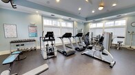 This fitness center offers a thoughtfully designed workout space that combines functionality with a bright, inviting atmosphere. The room is finished with soft blue-gray walls and crisp white trim, creating a calm and energizing environment conducive to exercise. Large windows with white plantation shutters line the walls, flooding the space with natural light while maintaining a clean, polished aesthetic. Recessed lighting and a pendant fixture overhead provide additional illumination throughout.

The facility is outfitted with a lineup of TREK commercial-grade cardio machines, including treadmills and ellipticals, arranged to maximize floor space and ease of use. A dedicated strength training area features a cable machine and an adjustable weight bench, while a dumbbell rack stocked with a range of free weights supports varied workout routines. A workout chart posted on the wall offers exercise guidance for users of all fitness levels.

The flooring is a durable rubber gym surface designed to absorb impact and support safe training. The overall layout is open and well-organized, allowing for comfortable movement between equipment. This fitness center delivers a complete exercise experience in a well-maintained, light-filled setting.