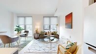 This thoughtfully designed single-floor unit offers an efficient and stylish layout, combining a kitchen and living room within one open, flowing space. The living area is bathed in natural light from a series of large floor-to-ceiling windows fitted with sleek roller shades, offering views of the surrounding urban streetscape. Light hardwood flooring runs throughout, complementing the bright white walls and creating a clean, modern aesthetic. The living space accommodates a comfortable seating arrangement anchored by a geometric-patterned area rug and a round glass coffee table with a brass frame, lending a contemporary and inviting atmosphere.

The kitchen is integrated seamlessly into the living area and features two-tone cabinetry — light grey upper cabinets and wood-grain lower cabinets — paired with white quartz countertops and a herringbone tile backsplash with under-cabinet lighting. Stainless steel appliances, including a range, built-in microwave, and dishwasher, complete the functional and refined kitchen.

The bathroom is finished with large-format neutral stone-look floor tiles, a sleek dark wood vanity with a white countertop, an illuminated mirror, and a full tub and shower combination with grid-pattern tile surround. A hallway connects the key areas of the unit, providing a logical and efficient flow throughout the space.