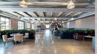 This expansive single-floor workspace masterfully blends industrial heritage with modern functionality. The open-plan layout provides generous room for a variety of work configurations, from collaborative desking areas to relaxed lounge seating, all flowing seamlessly across highly polished concrete floors that reflect the abundant natural light pouring in through large, multi-pane windows.

The architectural character of the space is defined by its exposed concrete coffered ceiling, complete with visible structural beams and industrial-style HVAC ductwork running overhead. Warm, original brick walls line the perimeter, lending a raw yet refined aesthetic that is further complemented by rows of pendant lighting suspended from the ceiling above. The entry foyer features stainless steel elevator doors set against the same exposed brick, reinforcing the building's industrial identity from the moment of arrival.

Throughout the main floor, lush green plants in large dark planters are strategically positioned to define zones and add warmth to the industrial palette. Dedicated workstation clusters with monitor setups are arranged alongside comfortable lounge seating groupings, including tufted sofas, upholstered chairs, and communal dining-height tables with leather barstools. A wall-mounted display screen anchors one end of the space, suitable for presentations or collaborative use. The overall ambiance is professional yet inviting, balancing productivity with comfort.