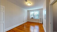 This single-floor unit is defined by warm, polished hardwood floors that run throughout the living areas and bedrooms, lending a cohesive and classic character to the space. The living area is generously proportioned, featuring neutral light gray walls, white baseboard trim, and a central flush-mount ceiling fixture. An electrical panel is mounted on one wall, and a doorway leads directly into the separate kitchen, which is equipped with dark espresso cabinetry, a black refrigerator, black microwave, and a white sink with a light-colored countertop, set atop vinyl flooring.

The bedrooms continue the hardwood flooring and neutral wall palette. Each room benefits from window-unit air conditioning and radiator heating. One bedroom features a built-in closet with double doors and a rod for hanging storage, while additional bedrooms offer windows that admit natural light and views of the surrounding exterior. The bathroom is finished with white subway tile wainscoting, mosaic tile flooring, a dark wood vanity with a white basin, a mirrored medicine cabinet, wall sconce lighting, and a full-size soaking tub with a shower rod and white tile surround. The overall ambiance is clean, functional, and classic.