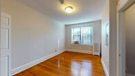 This single-floor unit is defined by warm, polished hardwood floors that run throughout the living areas and bedrooms, lending a cohesive and classic character to the space. The living area is generously proportioned, featuring neutral light gray walls, white baseboard trim, and a central flush-mount ceiling fixture. An electrical panel is mounted on one wall, and a doorway leads directly into the separate kitchen, which is equipped with dark espresso cabinetry, a black refrigerator, black microwave, and a white sink with a light-colored countertop, set atop vinyl flooring.

The bedrooms continue the hardwood flooring and neutral wall palette. Each room benefits from window-unit air conditioning and radiator heating. One bedroom features a built-in closet with double doors and a rod for hanging storage, while additional bedrooms offer windows that admit natural light and views of the surrounding exterior. The bathroom is finished with white subway tile wainscoting, mosaic tile flooring, a dark wood vanity with a white basin, a mirrored medicine cabinet, wall sconce lighting, and a full-size soaking tub with a shower rod and white tile surround. The overall ambiance is clean, functional, and classic.
