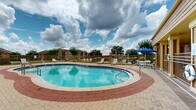 This outdoor recreational area centers around a generously sized freeform swimming pool with a classic kidney-inspired shape, finished with a blue-tiled interior that reflects clear, inviting water. The pool features convenient entry via built-in steps and safety handrails, with depth markers visible along the perimeter. Surrounding the pool is a spacious paved deck composed of decorative brick-pattern pavers, providing ample room for lounging and circulation. Numerous white lounge chairs and additional seating are distributed throughout the deck, accommodating a variety of outdoor activities. A shaded seating area, equipped with a table, chairs, and a large blue patio umbrella, offers a comfortable spot for relaxation out of direct sunlight. The pool area is fully enclosed by a metal safety fence, ensuring a secure environment. Adjacent to the pool, a covered clubhouse or amenity building with large windows and a pitched shingle roof provides an additional indoor-outdoor connection. A children's playground structure is visible beyond the fencing, extending the recreational offerings of the property. Mature trees and manicured landscaping line the perimeter, contributing to an open, airy atmosphere under expansive skies. The overall setting reflects a well-maintained, community-oriented outdoor amenity space.