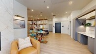 Located at 880 W 200 S in Salt Lake City, Utah, this well-appointed residential lobby offers a welcoming and thoughtfully designed communal space across a single floor. The layout is open and airy, with crisp white walls, recessed lighting, and light-toned hardwood flooring that runs throughout, lending a clean, contemporary aesthetic.

The lounge area serves as the focal point of the space, anchored by a striking floor-to-ceiling marble fireplace surround. Warm cognac leather armchairs are arranged around a low coffee table, creating an inviting seating vignette. Large floor-to-ceiling windows flood this area with natural light and provide views to the outdoors, enhancing the sense of openness.

A central seating zone features a plush, circular navy velvet sofa, adding a bold design statement to the middle of the space. Nearby, an open-frame wooden bookshelf serves as a stylish room divider, stocked with books and decorative objects that contribute to a curated, community-oriented atmosphere.

The mailroom area is well-equipped, featuring an extensive bank of black individual mailboxes lining the walls, complemented by a marble-topped central island for sorting packages. A gallery wall of framed landscape artwork adds warmth to this functional zone. Additionally, a modern automated package locker system is conveniently integrated into the space. Elevator access and secured entry doors are also present, completing this functional and design-forward lobby.