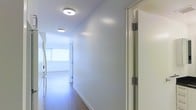 This single-floor space presents a thoughtfully designed interior centered around a long, well-lit hallway that connects the various areas of the home. The corridor is finished with warm-toned hardwood flooring that runs the full length of the space, creating a sense of continuity and flow throughout. Crisp white walls and clean white baseboards contribute to a bright, minimalist aesthetic that feels open and uncluttered.

Overhead, contemporary flush-mount ceiling lights are evenly spaced along the hallway, providing consistent and efficient illumination throughout. The lighting fixtures feature a modern, rounded profile that complements the clean lines of the space. A solid interior door with brushed metal hinges is visible along one side of the corridor, indicating well-defined, private room separations.

At the far end of the hallway, natural light filters in through a window fitted with layered roller blinds, softening the brightness while maintaining privacy. This natural light draws the eye forward and enhances the overall sense of depth and space. The overall ambiance is calm, modern, and functional, with a neutral palette that provides a versatile foundation for any interior style.