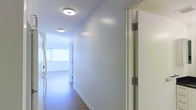 This single-floor space presents a thoughtfully designed interior centered around a long, well-lit hallway that connects the various areas of the home. The corridor is finished with warm-toned hardwood flooring that runs the full length of the space, creating a sense of continuity and flow throughout. Crisp white walls and clean white baseboards contribute to a bright, minimalist aesthetic that feels open and uncluttered.

Overhead, contemporary flush-mount ceiling lights are evenly spaced along the hallway, providing consistent and efficient illumination throughout. The lighting fixtures feature a modern, rounded profile that complements the clean lines of the space. A solid interior door with brushed metal hinges is visible along one side of the corridor, indicating well-defined, private room separations.

At the far end of the hallway, natural light filters in through a window fitted with layered roller blinds, softening the brightness while maintaining privacy. This natural light draws the eye forward and enhances the overall sense of depth and space. The overall ambiance is calm, modern, and functional, with a neutral palette that provides a versatile foundation for any interior style.