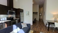 This single-floor space offers a thoughtfully designed open-concept layout that seamlessly blends the living, dining, and kitchen areas into one cohesive environment. The impressively tall ceilings lend a sense of openness and airiness throughout, making the space feel larger and more expansive than its footprint suggests.

The kitchen is outfitted with sleek, dark espresso cabinetry that contrasts beautifully against stainless steel appliances, including a full-size refrigerator, microwave, and range. The countertops and cabinetry are arranged efficiently to maximize functionality in the cooking area.

Rich hardwood-style flooring runs throughout the main living areas, adding warmth and continuity to the space. The living area comfortably accommodates a sofa and coffee table, bathed in natural light that highlights the neutral wall tones.

A long hallway leads toward the sleeping area, where a generous walk-in closet with built-in shelving and hanging rods provides ample storage. The overall color palette is clean and contemporary, with warm wood tones grounded by crisp white walls. Framed wall art adds subtle decorative touches throughout. The space is well-suited for those seeking a modern, efficiently designed environment with high-end finishes and practical storage solutions.