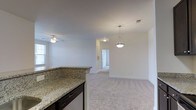 This single-floor residence offers a clean, modern aesthetic defined by an open-concept design that seamlessly connects the kitchen and living areas. The kitchen features a granite-topped peninsula with dark cabinetry, providing both a functional workspace and a natural dividing point between the cooking and living spaces. A stylish pendant light hangs above, adding a touch of elegance to the area.

The living space is finished with plush, light-toned carpeting that adds warmth and comfort underfoot, while a ceiling fan with an integrated light fixture promotes airflow and practicality. Crisp white walls and smooth ceilings enhance the sense of openness, allowing natural light from the window to brighten the interior. Recessed HVAC vents are integrated neatly into the ceiling, maintaining the clean lines of the space.

A hallway leads further into the residence, hinting at additional private rooms beyond. The layout flows naturally and feels spacious, making it well-suited for everyday living. Overall, the space strikes a balance between contemporary finishes and functional design, creating a comfortable and inviting environment throughout.