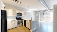 This well-appointed one-bedroom, one-bathroom unit offers a thoughtfully organized layout across a single floor, making efficient use of every square foot. The open-concept kitchen serves as the heart of the space, featuring white shaker-style cabinetry with open shelving, a peninsula with an undermount sink, stainless steel electric range, black appliances including a full-size refrigerator and built-in microwave, and light countertops. A pendant light and track lighting illuminate the workspace, while an exposed industrial duct adds a loft-like character to the design. Polished concrete floors extend through the kitchen and living area, contributing to the unit's modern, urban aesthetic. Large windows allow natural light to fill the living area, creating an open and airy feel. The bedroom is a quiet retreat finished with soft carpeting, light gray walls, and natural light from multiple windows fitted with blinds. A dedicated closet provides practical storage. The bathroom is cleanly appointed with a white vanity, undermount sink, mirrored cabinet, and a full tub-shower combination, maintaining the consistent clean, neutral palette found throughout the unit. Overall, the space presents a cohesive, contemporary design ideal for modern urban living.