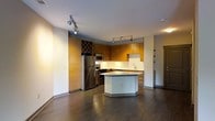 This single-floor space showcases a thoughtfully designed open-concept layout centered around a well-appointed kitchen. The kitchen features warm, medium-toned wood cabinetry that extends to the ceiling, providing generous storage throughout. A built-in diamond-pattern wine rack adds a distinctive architectural detail to the upper cabinetry, offering both functional storage and visual interest.

The kitchen is equipped with a full suite of stainless steel appliances, including a French door refrigerator, stainless range, and built-in microwave. A central island with a light grey countertop serves as both a workspace and casual gathering point, seamlessly connecting the kitchen to the living area. The perimeter countertops are finished in a clean white, complemented by a white tile backsplash that brightens the cooking area.

Track lighting installed on the ceiling provides directed illumination across both the kitchen and surrounding living space. Dark hardwood flooring runs throughout, creating a strong contrast against the white walls and light ceiling. The overall ambiance is modern and functional, with a clean, neutral palette accented by the richness of the wood cabinetry and flooring.