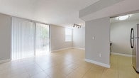 This single-floor residence is designed with a clean, contemporary aesthetic throughout. Light-toned ceramic tile flooring runs consistently across all living areas, complementing the neutral gray walls and white baseboards to create a bright, cohesive interior.

The kitchen is a functional centerpiece, featuring a pass-through peninsula topped with granite countertops that overlooks the main living space. White shaker-style cabinetry lines the walls, paired with stainless steel appliances including a range, microwave, refrigerator, and dishwasher. A double stainless steel sink with a modern pull-down faucet adds practicality to the well-appointed workspace.

The living area is generously proportioned and filled with natural light, courtesy of a large sliding glass door fitted with vertical blinds that opens to an outdoor area. An additional window further brightens the space, which also features track lighting overhead and a textured ceiling throughout.

A hallway leads to a spacious bedroom with direct access to a private bathroom. The bathroom is finished in crisp white, featuring a full-size soaking tub with tiled surround. The vanity area includes white cabinetry and granite countertops, maintaining consistency with the kitchen's design language.