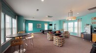 This single-floor community clubhouse is designed to offer a variety of functional and recreational spaces. The main lounge and game area is the centerpiece of the facility, featuring bold teal-painted walls accented by a deep red feature wall that creates a lively, energetic atmosphere. The space is generously sized, accommodating a full pool table, multiple seating arrangements including a leather sofa, patterned accent chairs, and a round coffee table. A wall-mounted flat-screen television provides entertainment, while pendant and drum-shade light fixtures add warmth overhead. Multiple windows along the perimeter flood the room with natural light and offer views of the surrounding exterior. High-top bar tables and stools create an additional casual seating zone within the lounge. A dedicated business center is located in an adjacent room, painted in a warm orange tone. It features multiple individual workstations equipped with desktop computers, ergonomic chairs, and a shared printer. Built-in cabinetry provides additional storage, and decorative metallic wall art adds visual interest. Windows in this room ensure a well-lit work environment. Carpeted flooring runs throughout the facility, contributing to a comfortable and cohesive feel across all areas.