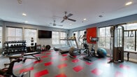 Located at 750 Summer St in Stamford, Connecticut, this fitness center is a well-appointed, single-level workout space designed to support a full range of exercise routines. The room features a striking grey and red tile floor that adds a bold, energetic aesthetic throughout. Dual ceiling fans provide consistent airflow and comfort during workouts, complemented by recessed lighting that keeps the space bright and functional.

Multiple windows line the walls, inviting generous natural light and offering views of the surrounding neighborhood, creating an open and motivating atmosphere. A large wall-mounted flat-screen television serves the cardio zone, where treadmills, an elliptical trainer, and a stationary bike are neatly arranged along the windowed wall.

The free weights section is thoughtfully organized, featuring a full rack of dumbbells, kettlebells, a weight bench, and medicine balls, all positioned alongside a floor-to-ceiling mirror that enhances the sense of space. Additional equipment includes a multi-functional cable machine and cycling bikes for a comprehensive training experience.

Motivational signage adorns the walls, reinforcing the gym's energizing character. The space is efficiently laid out to maximize usability while maintaining clear circulation throughout, making it a practical and well-equipped facility for a wide variety of fitness activities.