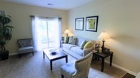 This single-floor space offers a welcoming and thoughtfully designed living environment. The living room serves as the heart of the home, bathed in natural light streaming through large sliding glass doors that open to a private balcony, offering a seamless connection to the outdoors. Neutral, warm-toned walls provide a calm backdrop that complements the plush carpeted flooring underfoot.

The living room is furnished with a classic sofa adorned with green and patterned accent pillows, paired with an elegant upholstered armchair featuring refined nail-head trim detailing. A dark-finished rectangular coffee table anchors the seating area, while matching end tables with table lamps add a warm, ambient glow to the space. Framed botanical artwork in bold black frames adds a stylish focal point to the wall above the sofa.

Sheer curtains frame the sliding glass doors, softening the natural light and enhancing the airy feel of the room. The overall ambiance is one of understated elegance — casual yet polished, with a cohesive color palette of neutrals, greens, and dark wood tones that create a harmonious and inviting atmosphere throughout the space.