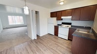 This single-floor, two-bedroom, one-bathroom unit offers a practical and well-organized layout across a comfortably sized floor plan. The kitchen is a functional L-shaped space equipped with rich dark brown cabinetry, dark countertops, and white appliances including a refrigerator, electric range, and under-cabinet range hood. Light wood-style vinyl flooring runs throughout the kitchen, which also features a window that brings in natural light. A decorative pendant light and a closet positioned just off the kitchen add both character and convenient storage to the area.

The living room is generously proportioned, featuring neutral wall-to-wall carpeting, bright white walls, and a large multi-pane window that allows ample natural light to fill the space. Baseboard heating runs along the exterior walls throughout the unit.

Both bedrooms are carpeted and offer a clean, neutral palette. Each room includes a window with outdoor views and a dedicated closet with shelving. One bedroom also features a wall-mounted heating and cooling unit for individual climate control.

The bathroom is finished with wood-style vinyl flooring, a white vanity with a light countertop, a tiled tub-shower combination, and a baseboard heater, completing this straightforward and functional layout.