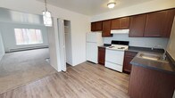 This single-floor, two-bedroom, one-bathroom unit offers a practical and well-organized layout across a comfortably sized floor plan. The kitchen is a functional L-shaped space equipped with rich dark brown cabinetry, dark countertops, and white appliances including a refrigerator, electric range, and under-cabinet range hood. Light wood-style vinyl flooring runs throughout the kitchen, which also features a window that brings in natural light. A decorative pendant light and a closet positioned just off the kitchen add both character and convenient storage to the area.

The living room is generously proportioned, featuring neutral wall-to-wall carpeting, bright white walls, and a large multi-pane window that allows ample natural light to fill the space. Baseboard heating runs along the exterior walls throughout the unit.

Both bedrooms are carpeted and offer a clean, neutral palette. Each room includes a window with outdoor views and a dedicated closet with shelving. One bedroom also features a wall-mounted heating and cooling unit for individual climate control.

The bathroom is finished with wood-style vinyl flooring, a white vanity with a light countertop, a tiled tub-shower combination, and a baseboard heater, completing this straightforward and functional layout.