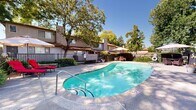 Located at 109 Herman Cir in Auburn, California, this outdoor amenity area serves as a central gathering space within a well-established residential community. The centerpiece of the space is a generously sized, kidney-shaped swimming pool with clear turquoise water, accessible via built-in entry steps and metal handrails. The pool is surrounded by a spacious concrete deck that provides ample room for lounging and movement.

The poolside area is furnished with comfortable seating options, including vibrant red lounge chairs and a shaded patio table set beneath a large umbrella, ideal for relaxing in the warm Northern California climate. The entire pool area is enclosed by a black wrought-iron security fence with a gated entry, ensuring safety and controlled access. Signage within the enclosure provides pool rules and capacity information.

Mature, leafy trees frame the perimeter of the space, offering natural shade and a lush, park-like atmosphere under clear blue skies. Manicured hedges and potted plants add greenery and visual appeal throughout. The surrounding two-story residential buildings with stucco exteriors and pitched rooflines are visible beyond the fence, providing context to the community setting. The overall ambiance is relaxed, bright, and inviting, well-suited for outdoor recreation and leisure.