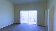 This single-floor space presents a straightforward, functional layout centered around a carpeted living area. The room is finished with neutral, light yellow walls that create a clean and understated backdrop. A prominent feature of the space is the large sliding glass door, which spans a generous portion of one wall and is fitted with vertical blinds. These blinds offer adjustable light control while still allowing natural light to filter through, brightening the interior. The sliding door provides a direct connection to the outdoors, with a view of an exterior structure visible through the glass. The wall-to-wall carpeting in a muted gray-blue tone adds a layer of comfort underfoot and contributes to the room's quiet, neutral aesthetic. The ceiling is smooth and unadorned, maintaining a simple, clean look throughout. The overall ambiance of the space is modest and move-in ready, offering a blank canvas well-suited for personalization. The combination of natural light from the large glass door and the neutral color palette gives the room an open, airy feel despite its unpretentious design.