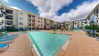 The Cool Springs Oasis Pool is a expansive resort-style outdoor amenity nestled within a four-story residential community. The centerpiece of this courtyard space is a large, multi-section swimming pool with clear turquoise water, featuring a striking decorative stone fountain as a focal point. The pool spans multiple interconnected areas, including a shallower wading section and a broader lap-friendly zone, with depth markers clearly visible along the edges. Surrounding the pool, a spacious paved deck finished in warm terracotta-toned pavers provides ample room for relaxation, lined with vibrant blue lounge chairs positioned to maximize sun exposure. Raised planting beds and lush landscaping with palm trees, ornamental grasses, and manicured shrubs add a tropical ambiance throughout the courtyard. Patio umbrellas offer shaded seating areas for residents seeking refuge from the sun. Well-maintained green lawn areas and additional garden beds are interspersed around the perimeter walkways. The four-story residential buildings with navy blue awnings and private balconies frame the courtyard on all sides, creating an enclosed, private retreat. Decorative street lamps illuminate the paved walkways, enhancing the space's functionality and aesthetic appeal day and night.