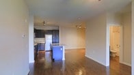 This single-floor space showcases an open-concept layout that seamlessly connects the kitchen and living area, creating a spacious and functional environment. The rich, dark hardwood floors extend throughout, adding warmth and a polished aesthetic to the interior.

The kitchen is well-appointed with dark cabinetry that provides ample storage, light-toned countertops, and stainless steel appliances, including a refrigerator and range. A central island with a contrasting blue base and light countertop serves as a natural dividing point between the kitchen and the open living area, offering additional workspace or casual seating opportunities.

Track lighting is installed on the ceiling in both the kitchen and living areas, ensuring even, adjustable illumination throughout the space. The living area is generously sized with neutral-toned walls that complement the flooring and create a versatile backdrop for various interior styles.

The open floor plan promotes an airy, connected feel, making the space ideal for both everyday living and entertaining. Clean baseboards and a straightforward layout contribute to the overall tidy and functional character of this well-designed single-floor residence.