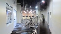 This fitness facility offers a thoughtfully designed workout environment that combines industrial aesthetics with modern functionality. The space features a striking contrast of design elements, including exposed brick walls, a dark ceiling with visible ductwork, and bright track lighting that illuminates the floor throughout. The flooring transitions between a patterned, multi-toned carpet in the main workout area and a dark hardwood-style surface along the perimeter, adding visual depth to the layout. The cardio zone is well-stocked with a row of treadmills, ellipticals, and upright exercise bikes, all positioned to allow ample space between machines. Strength training equipment, including cable machines and resistance apparatuses, occupies the central floor space and is arranged to maximize usability and flow. The perimeter walls feature motivational signage and decorative touches such as tall potted plants, contributing to an energizing atmosphere. Mirrored walls enhance the sense of openness and allow users to monitor their form during workouts. The high ceilings and open layout create a spacious, airy feel, making this a comprehensive and inviting facility suited for a wide range of fitness activities.