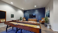 Located at 73 Brownstone Way in Englewood, NJ, this single-floor community lounge is designed to offer residents a well-rounded social and recreational experience. The space is thoughtfully laid out to accommodate both lively gatherings and relaxed downtime.

At the heart of the lounge, a full-size shuffleboard table commands attention, anchored atop a bold blue geometric-patterned area rug that adds a striking visual element to the hardwood-style flooring throughout. Facing the shuffleboard is a comfortable leather sofa accompanied by a marble-top coffee table, creating an inviting seating area ideal for casual conversation or relaxation.

A deep navy blue accent wall serves as a dramatic focal point, featuring a wall-mounted flat-screen television flanked by warm cognac leather wingback chairs and a wooden media console. Potted greenery is distributed throughout the space, adding natural freshness to the contemporary interior.

A dedicated game corner showcases a large wall-mounted Scrabble board alongside a compact bistro-style table with stools, while a collection of board games is neatly stacked nearby, encouraging interactive play. Large windows along one wall allow natural light to flow in, offering views of the surrounding brick exterior streetscape.

Black-framed glass doors provide a modern architectural detail and connect the lounge to adjacent areas, enhancing the open, accessible feel of the space.