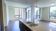 This single-floor residence offers a thoughtfully designed open-concept layout that seamlessly blends the kitchen and living areas into one inviting, light-filled space. The kitchen is a standout feature, anchored by a generous island topped with polished granite countertops and a deep double-basin stainless steel sink. Dark espresso cabinetry provides rich contrast against the lighter stone surfaces, while two elegant pendant lights hang above the island, adding warmth and style to the workspace. The open living area extends naturally from the kitchen, finished with rich hardwood flooring that runs throughout the space. Floor-to-ceiling windows line multiple walls, flooding the interior with abundant natural light and offering views of surrounding greenery and the neighboring urban streetscape. The large window panels are framed in sleek black trim, adding a contemporary architectural touch that complements the modern aesthetic of the home. The elevated ceiling height further enhances the sense of openness and volume. The overall ambiance is clean, contemporary, and welcoming, making it an ideal space for both comfortable everyday living and entertaining.