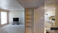 This single-floor space offers a practical and straightforward layout centered around an open living area. A notable feature is the built-in shelving unit integrated into a dividing wall, providing convenient storage or display space across multiple shelves. The living area is carpeted in a neutral tone, contributing to a warm and understated ambiance. A front entry door is visible in the background, suggesting the living space flows naturally from the main entrance. The walls are finished in a soft, neutral palette, and the textured ceiling adds a subtle architectural detail throughout. The open sightlines between areas create a connected feel, making the space feel functional and cohesive. Overall, the layout is simple and efficient, with built-in storage serving as a practical centerpiece.