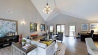 This single-story clubhouse is defined by its dramatic vaulted ceilings, which rise to an impressive peak and lend the interior a grand, open atmosphere. A statement chandelier with multiple globe lights anchors the central living area, serving as a focal point that draws the eye upward and enhances the sense of vertical space. The walls are finished in a clean, neutral gray palette, complemented by crisp white ceiling beams that add architectural character throughout.

The main gathering area is thoughtfully arranged with plush white sofas, patterned accent chairs in gray and blue tones, and a glass-topped coffee table, creating an inviting space for socializing. Decorative mirrors with gray frames line one wall, adding depth and reflecting light throughout the room. Light hardwood-style flooring runs throughout, contributing warmth and continuity.

Large glass doors and windows along the far wall flood the space with natural light while offering views of the surrounding outdoor greenery. A secondary seating and dining area is visible near the windows, providing versatile options for gatherings. The overall ambiance is modern and polished, balancing comfort with refined design.