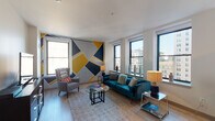 This stylish single-floor urban residence makes a strong design statement through its bold geometric accent walls, rendered in a striking palette of navy, gold, and gray. These eye-catching murals unify the living and bedroom spaces with a cohesive artistic identity.

The spacious living room is anchored by multiple large windows that frame impressive city skyline views, flooding the room with natural light. A plush teal velvet sofa, a glass-top coffee table, and a woven area rug complete the contemporary aesthetic, while light hardwood flooring extends throughout the main living areas.

The kitchen is thoughtfully outfitted with dark espresso cabinetry, white countertops, white subway tile backsplash, and a full suite of stainless steel appliances, including a gas range, microwave, and a large French-door refrigerator. Track lighting overhead ensures excellent task illumination.

Two bedrooms offer comfortable retreats; one features a vibrant golden-yellow accent wall with a window overlooking neighboring architecture, while the other carries the geometric mural theme and includes a double-door closet. Both rooms are carpeted for warmth and comfort.

The bathroom is clean and modern, finished with light tile flooring, a tub-shower combination with white subway tile surround, and a sleek vanity with white countertop. The overall ambiance of the space is energetic yet livable, blending urban sophistication with bold personal style.