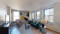 This stylish single-floor urban residence makes a strong design statement through its bold geometric accent walls, rendered in a striking palette of navy, gold, and gray. These eye-catching murals unify the living and bedroom spaces with a cohesive artistic identity.

The spacious living room is anchored by multiple large windows that frame impressive city skyline views, flooding the room with natural light. A plush teal velvet sofa, a glass-top coffee table, and a woven area rug complete the contemporary aesthetic, while light hardwood flooring extends throughout the main living areas.

The kitchen is thoughtfully outfitted with dark espresso cabinetry, white countertops, white subway tile backsplash, and a full suite of stainless steel appliances, including a gas range, microwave, and a large French-door refrigerator. Track lighting overhead ensures excellent task illumination.

Two bedrooms offer comfortable retreats; one features a vibrant golden-yellow accent wall with a window overlooking neighboring architecture, while the other carries the geometric mural theme and includes a double-door closet. Both rooms are carpeted for warmth and comfort.

The bathroom is clean and modern, finished with light tile flooring, a tub-shower combination with white subway tile surround, and a sleek vanity with white countertop. The overall ambiance of the space is energetic yet livable, blending urban sophistication with bold personal style.
