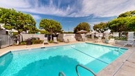 Located at 301-307 Avocado St in Costa Mesa, California, this outdoor communal pool area offers a welcoming retreat designed for relaxation and leisure under the Southern California sun. The centerpiece is a generously sized rectangular swimming pool with clear, blue water, framed by a clean concrete pool deck that provides ample space for sunbathing and socializing.

The pool area is well-appointed with multiple white lounge chairs and a shaded seating arrangement featuring a patio umbrella, table, and chairs — ideal for those seeking a break from the sun. Colorful potted flowering plants, including vibrant red blooms, are thoughtfully placed around the perimeter, adding a welcoming touch to the environment.

The space is enclosed by a combination of metal fencing and a low brick-style perimeter wall, providing both security and a sense of privacy. Mature trees surround the area, offering natural shade and enhancing the lush, park-like atmosphere. The adjacent multi-story building features a distinctive design with teal-accented awnings and private balconies that overlook the pool courtyard, reinforcing the resort-like character of the property. The well-maintained landscaping with green lawns visible beyond the pool fence further contributes to the inviting, serene ambiance of this outdoor amenity space.