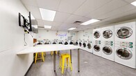 Located at 800 N Pennsylvania St in Denver, Colorado, The Missing Sock is a clean and modern laundry facility designed for efficiency and comfort. The single-floor space is thoughtfully laid out, with rows of stacked and standalone front-loading washers and dryers lining the perimeter walls. The machines are uniformly white with chrome detailing, contributing to the bright, orderly aesthetic of the room. Large glass partition windows divide sections of the facility, allowing natural light to flow through and maintaining an open, airy feel beneath the drop-panel ceiling with recessed lighting. The flooring throughout is a durable gray tile, practical for a high-traffic environment. A central waiting and folding area features white tables with vibrant yellow metal stools, adding a cheerful pop of color to the space. A wall-mounted flat-screen TV provides entertainment for guests during their visit. Safety essentials, including a fire extinguisher and hand sanitizer dispenser, are clearly visible on the walls, alongside posted occupancy guidelines. A neon sign reading "The Missing Sock" adorns one wall, giving the facility a welcoming personality. The overall ambiance is clean, well-lit, and functional, making it a practical and pleasant destination for laundry needs in the heart of Denver.
