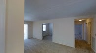 This single-floor residential space offers a straightforward and functional layout designed for comfortable everyday living. The main living area is finished with wall-to-wall carpeting in a neutral beige tone, creating a warm and cohesive feel throughout. Smooth, light-colored walls paired with a clean ceiling contribute to a bright and airy atmosphere. Natural light filters in through multiple windows, including a large window or sliding door along one wall that enhances the sense of openness. A doorway leads from the main living area into an adjoining room, which also features carpeted flooring and its own window, allowing for continued natural illumination. The connecting layout between rooms promotes an easy flow through the space. Electrical outlets are positioned throughout, and the walls are freshly finished in a soft white tone, lending the space a clean and neutral canvas suitable for a variety of interior styles. The overall ambiance is modest and practical, with an uncluttered design that emphasizes usability and comfort. The space presents itself in a well-maintained, move-in ready condition, ideal for those seeking a simple and welcoming residential environment.