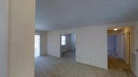 This single-floor residential space offers a straightforward and functional layout designed for comfortable everyday living. The main living area is finished with wall-to-wall carpeting in a neutral beige tone, creating a warm and cohesive feel throughout. Smooth, light-colored walls paired with a clean ceiling contribute to a bright and airy atmosphere. Natural light filters in through multiple windows, including a large window or sliding door along one wall that enhances the sense of openness. A doorway leads from the main living area into an adjoining room, which also features carpeted flooring and its own window, allowing for continued natural illumination. The connecting layout between rooms promotes an easy flow through the space. Electrical outlets are positioned throughout, and the walls are freshly finished in a soft white tone, lending the space a clean and neutral canvas suitable for a variety of interior styles. The overall ambiance is modest and practical, with an uncluttered design that emphasizes usability and comfort. The space presents itself in a well-maintained, move-in ready condition, ideal for those seeking a simple and welcoming residential environment.