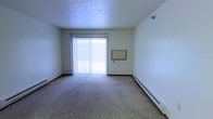 This single-floor space offers a clean, functional layout designed for comfortable everyday living. The main living area is finished with wall-to-wall carpet that adds warmth underfoot, while a textured ceiling runs throughout, giving the space a classic residential character. A large sliding glass door, dressed with vertical blinds, serves as a prominent architectural feature, flooding the room with natural light and providing direct access to an outdoor balcony — offering a seamless indoor-outdoor connection. The walls are painted in a crisp, neutral white, creating a bright and open atmosphere that makes the space feel airy and versatile. A baseboard heating system runs along the exterior wall, ensuring efficient climate control during cooler months. A wall-mounted panel, likely an intercom or thermostat, is also present, adding to the unit's practical amenities. The overall ambiance is simple and understated, presenting a blank canvas well-suited for a variety of furnishing styles and personal touches.