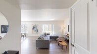 This single-floor residence is designed with a clean, modern aesthetic throughout, characterized by light-toned walls, light wood-style flooring, and an abundance of natural light. The spacious living room serves as the centerpiece of the home, featuring a sliding glass door that opens to an outdoor area with green views, vaulted ceiling sections, and ample room for comfortable seating arrangements. A round wall mirror and contemporary décor accents contribute to the room's polished, airy feel.

The kitchen is well-appointed with white shaker-style cabinetry, dark countertops, a tile backsplash, stainless steel appliances including a range, microwave, and refrigerator, and a deep basin sink. A peninsula with bar stool seating provides a casual dining option and connects the kitchen to the main living space.

A dedicated dining nook offers a window with outdoor views and pendant lighting overhead. The bedroom is quietly appointed with a reach-in closet featuring louvered bifold doors. The full bathroom includes a tub-shower combination with classic white subway tile, a white vanity, and stone-style tile flooring, completing the home's consistently clean and contemporary interior palette.
