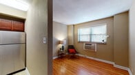 This single-floor unit offers a well-organized layout with consistent design elements throughout. Warm honey-toned hardwood flooring flows through the main living and sleeping areas, creating a cohesive and inviting atmosphere, while the kitchen and bathroom feature lighter tile flooring. The walls are finished in a neutral taupe tone, complemented by clean white baseboards and a textured ceiling.

The main living area receives natural light through a window fitted with horizontal blinds, and a wall-mounted PTAC unit provides climate control. A partial wall with a textured finish defines the entry zone, which includes a coat closet with a full-width hanging rod. The closet is accessible via double doors and sits adjacent to the unit's entry door.

A second interior room, visible from the main space, features a white panel door and connects to the rest of the layout via an open hallway. The kitchen is efficiently designed with rich cherry-toned cabinetry, granite-look countertops, stainless steel appliances including a refrigerator, over-the-range microwave, and electric range. The bathroom, glimpsed from the living area, includes a shower with a curtain, toilet, and a vanity with dark cabinetry.