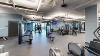 Located at 1803 S 46th St in Rogers, Arkansas, this fitness facility offers a generously sized single-floor layout designed to accommodate a full range of workout needs. The open floor plan is organized into distinct functional zones, allowing for efficient use of the space without feeling cramped.

The cardio zone features a robust lineup of treadmills, ellipticals, stair climbers, and rowing machines, all positioned to maximize accessibility and flow. Adjacent to this area, the strength training zone is outfitted with a comprehensive selection of cable machines, plate-loaded equipment, resistance machines, and free weights including dumbbells and benches — providing options for users of all fitness levels.

Throughout the facility, durable rubber flooring provides traction and impact absorption, while large wall-mounted mirrors enhance the sense of openness and allow users to monitor their form. Overhead, exposed ductwork and piping run along the ceiling, lending the space an industrial aesthetic that is complemented by soft blue accent walls and built-in shelving units. Linear LED lighting ensures the entire floor is evenly and brightly illuminated.

Ceiling fans are strategically placed to support air circulation, and mounted flat-screen televisions are visible throughout the space. A glazed glass partition wall adds a modern architectural touch, while windows along one end allow natural light and outdoor views to filter in, contributing to an energizing and welcoming workout environment.