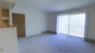 This single-floor space features an open living area finished with wall-to-wall carpet in a neutral tone, contributing to a clean and functional atmosphere. The room is defined by smooth white walls and a flat ceiling, creating a simple, uncluttered aesthetic throughout. A large sliding glass door dominates one wall, fitted with vertical blinds that allow for adjustable natural light and outdoor views, brightening the interior considerably. Along the base of the exterior wall, a baseboard heating unit provides efficient climate control. A wooden interior door is positioned along the adjacent wall, offering access to other areas of the space. Standard electrical outlets and light switch plates are positioned at conventional heights throughout the room. The overall layout is straightforward and open, providing flexibility for a variety of furniture arrangements. The neutral finishes — including the light carpet, white walls, and minimal trim details — lend the space a versatile, move-in-ready quality suitable for a range of uses.
