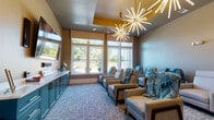 Located at 4101 Glenloch Creek in Matthews, NC, this single-floor media and lounge space combines contemporary design with comfort-focused amenities. The room is anchored by a striking pair of starburst chandeliers that cast warm, dramatic light throughout the space, complementing a ceiling detail finished in a rich gold tone.

Seating is plentiful and varied, featuring rows of plush reclining chairs draped with cozy teal faux-fur throws, ideal for relaxed viewing. A built-in nook along one wall offers a distinct lounge vibe, with a vibrant red bench seat, bold multicolored patterned upholstery, yellow accent pillows, and petite round side tables — creating an inviting social corner.

A large flat-screen television is mounted on the wall above a teal cabinetry unit with brass hardware, providing media storage and surface space. Adjacent to the screen, a classic popcorn machine adds a fun, cinematic touch. Three bold, colorful framed art pieces line the wall nearby, contributing to the room's energetic aesthetic.

Large windows along one wall flood the space with natural light and offer views of the surrounding greenery and streetscape, while patterned woven valances add a decorative finish. The room is carpeted in a multi-toned blue-grey textile that ties the color palette together, creating a cohesive and inviting atmosphere throughout.
