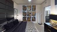 This single-floor residence showcases a cohesive, boldly styled interior that balances contemporary design with eclectic artistic flair. The living area centers around a tufted leather sofa adorned with deep navy velvet pillows, complemented by a sculptural hand-shaped accent chair and a geometric patterned area rug. Plaid navy-and-gold drapes frame a full-height sliding glass door that opens to an outdoor patio, allowing natural light to filter into the space. A compact desk area with a gold-framed mirror adds a functional yet decorative touch.

The kitchen is outfitted with two-tone cabinetry—crisp white uppers paired with dark lower cabinets—set against a mosaic tile backsplash. Stainless steel appliances, including a refrigerator and dishwasher, and dark granite countertops complete the polished, modern aesthetic.

The bedroom features a dramatic floor-to-ceiling upholstered black headboard flanked by framed black-and-white artwork and matching white nightstands with brass accents. A bold nine-panel artwork installation decorates an adjacent wall, reinforcing the room's curated, gallery-like atmosphere. Carpet flooring adds warmth underfoot.

The bathroom includes a tub-shower combination with a boldly patterned curtain, dark vanity cabinetry with granite countertops, wood-look flooring, and direct access to a walk-in closet with wire shelving.