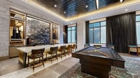 Located at 1000 Broadway in the heart of Nashville, TN, this elegantly appointed game room offers a premium entertainment experience across a generously sized single-floor layout. The space is defined by refined finishes throughout, including large-format stone tile flooring, textured stone accent walls, and a striking dark, metallic ceiling with warm cove lighting that creates a sophisticated ambiance.

At the heart of the space sits a full wet bar with a marble countertop, leather-upholstered bar stools with brass accents, and an iridescent tile backsplash framed by custom built-in shelving — a true focal point for social gatherings. A regulation-sized billiards table anchors one area, while a shuffleboard table, foosball table, and a central lounge area with a round dining table and wicker chairs provide diverse entertainment options.

A wall-mounted flat-screen television above a sleek console creates a comfortable media viewing area, complemented by curated artwork displayed on crisp white walls. Large floor-to-ceiling windows with plaid drape panels flood the space with natural light and offer views of the surrounding Nashville cityscape. An access point leads to an outdoor terrace, further expanding the entertainment possibilities. Oversized spherical pendant lights add a bold, modern design statement throughout.