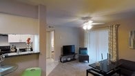 This single-story space showcases a functional and straightforward layout centered around an open living area. The living room features a combination of tile and carpet flooring, providing a practical transition between spaces. A white ceiling fan with a built-in light fixture serves as a central overhead feature, ensuring both airflow and ambient lighting throughout the room. Textured ceilings add a subtle architectural detail overhead.

Large sliding glass doors fitted with vertical blinds allow natural light to fill the living area while offering the option for privacy. The neutral gray wall tones create a calm, understated backdrop throughout. A partial wall with a countertop bar overhang separates the living space from an adjacent kitchen area, visible to the left, which features dark granite-style countertops and tile flooring.

A hallway visible from the main living area leads to additional rooms, suggesting a well-connected interior flow. The overall ambiance is modest and practical, with a layout designed to maximize usability within a compact footprint. The combination of tile and carpet flooring throughout the space balances comfort and easy maintenance.