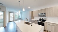 This well-appointed one-bedroom, one-bathroom unit offers a thoughtfully designed open-concept layout across a single floor. The kitchen is a standout feature, showcasing light wood flat-front cabinetry extending to the ceiling, a white quartz countertop island with an undermount sink and brushed gold faucet, pendant lighting, and a full suite of stainless steel appliances including a French door refrigerator, electric range, and over-the-range microwave. A white subway tile backsplash adds a clean, contemporary touch. Warm-toned wood-look flooring flows throughout the main living areas, creating a cohesive and inviting aesthetic. The living room is bathed in natural light through large windows and a sliding glass door that frame views of lush greenery and a pool, with a ceiling fan adding comfort. The bedroom features plush neutral carpet, a ceiling fan, and a bright window with outdoor views, providing a calm retreat. The bathroom is finished with large-format tile flooring, a light wood vanity with quartz countertop, a dark-framed mirror, a tall storage tower, and ample drawer space. Clean white walls throughout contribute to an airy, modern ambiance that is both functional and stylish.