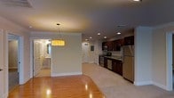 This single-floor residence offers an open-concept layout that seamlessly connects the living, dining, and kitchen areas, creating a bright and functional living environment. The kitchen is equipped with dark espresso cabinetry, stainless steel appliances including a refrigerator, range, and microwave, and light-toned countertops that provide a clean contrast to the rich cabinetry. Recessed ceiling lights illuminate the kitchen and main living area, while a warm, amber pendant light fixture adds a decorative focal point above the dining space. Crown molding lines the upper edges of the walls, lending a refined architectural detail to the interior. The flooring transitions between polished hardwood in the living and dining areas and a neutral tile in the kitchen zone, offering both warmth and practicality. A glimpse into the bathroom reveals a round mirror, vanity, and bright lighting, suggesting a well-designed and functional space. Neutral-toned walls throughout contribute to an airy, cohesive ambiance. The layout flows naturally from room to room, making efficient use of the available space while maintaining an open and inviting atmosphere.