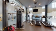 Located in the heart of downtown Winston-Salem, NC, this well-appointed fitness center offers a comprehensive workout environment designed to accommodate a variety of exercise routines. The space is defined by its modern aesthetic, featuring floor-to-ceiling frosted glass partitions with bold striped detailing that divide the room into distinct functional zones while maintaining an open, airy feel. Natural light filters in through windows positioned along the upper walls, complementing the recessed ceiling lighting for a bright, energizing atmosphere. Durable rubberized flooring provides safety and comfort throughout the workout areas, while a classic checkered tile floor is visible in the adjoining section beyond the glass dividers. The cardio zone is well-equipped with multiple treadmills and elliptical machines, all positioned beneath a ceiling-mounted flat-screen television for entertainment during workouts. The strength training area features a robust cable and pulley machine system with rope attachments, as well as a full rack of dumbbells and an adjustable workout bench for targeted exercises. Stability balls round out the functional fitness offerings. The space benefits from a clean, contemporary design that creates a professional and motivating environment, making it a fully equipped destination for both cardiovascular and strength training needs.