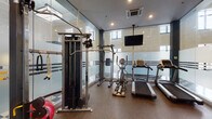 Located in the heart of downtown Winston-Salem, NC, this well-appointed fitness center offers a comprehensive workout environment designed to accommodate a variety of exercise routines. The space is defined by its modern aesthetic, featuring floor-to-ceiling frosted glass partitions with bold striped detailing that divide the room into distinct functional zones while maintaining an open, airy feel. Natural light filters in through windows positioned along the upper walls, complementing the recessed ceiling lighting for a bright, energizing atmosphere. Durable rubberized flooring provides safety and comfort throughout the workout areas, while a classic checkered tile floor is visible in the adjoining section beyond the glass dividers. The cardio zone is well-equipped with multiple treadmills and elliptical machines, all positioned beneath a ceiling-mounted flat-screen television for entertainment during workouts. The strength training area features a robust cable and pulley machine system with rope attachments, as well as a full rack of dumbbells and an adjustable workout bench for targeted exercises. Stability balls round out the functional fitness offerings. The space benefits from a clean, contemporary design that creates a professional and motivating environment, making it a fully equipped destination for both cardiovascular and strength training needs.