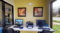 This business center is a compact yet fully functional workspace designed to meet professional needs. The room features a long, built-in desk running along one wall, outfitted with two desktop computer workstations complete with monitors, keyboards, and mice, as well as a centrally positioned multifunction printer and scanner for convenient shared use. Ergonomic mesh-back chairs provide comfortable seating at each workstation.

The space is finished with warm yellow walls that lend an inviting, energetic atmosphere, accented by two bold, framed abstract artworks in rich red and orange tones displayed above the desk. Wood-look flooring adds a clean, modern touch to the room.

A standout feature of this business center is the expansive floor-to-ceiling window that floods the space with natural light and offers a pleasant view of the well-maintained landscaping outside, including colorful flower beds and manicured greenery. A glass-panel door provides additional visual connection to the exterior covered entrance, which features classic brick columns and a welcoming entry mat. A private interior door also connects the center to adjacent areas of the building, ensuring easy accessibility and flow throughout the facility.