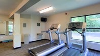 This fitness center provides a focused and functional environment designed for cardiovascular exercise and general wellness. The space is outfitted with multiple treadmills positioned to take advantage of a window overlooking a lush green outdoor area, allowing natural light to filter in and providing an pleasant view during workouts. The room features durable carpeted flooring, neutral-toned walls, and a drop ceiling with overhead fluorescent lighting that ensures the space is well-lit throughout. A wall-mounted flat-screen television is positioned at eye level for convenient viewing during exercise sessions. A built-in water fountain is conveniently located along the wall, offering easy access to hydration. Safety and compliance features, including a fire alarm, are visibly installed. An electrical panel box on the wall reflects the utilitarian, practical nature of the space. The overall ambiance is clean, straightforward, and purpose-driven, making it well-suited as an amenity fitness room, such as those found in hotels, residential communities, or office complexes. The layout is efficient, maximizing the available floor area to accommodate equipment while maintaining clear circulation paths.